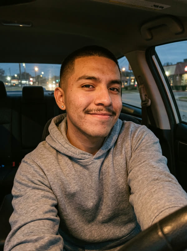 Front-camera selfie of a young man with a thin mustache in a grey hoodie, taken in a parked car