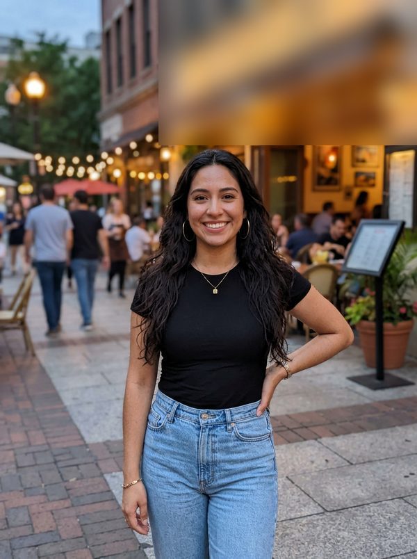Evening street photo of a smiling young woman with long curly hair, gold hoop earrings, a black tee and jeans